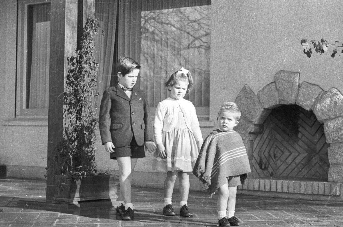 Plano entero de dos niños y un niña parados de mayor a menos estatura. La Niña se encuentra parada en el medio de la fila. El niño de menor edad lleva puesta una manta. Se encuentran ubicados en el patio de una casa durante la tarde