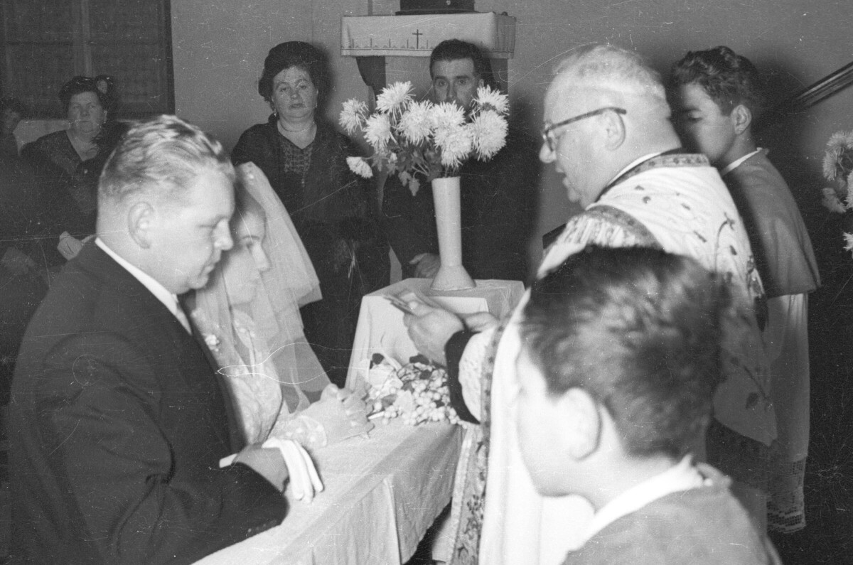 Plano medio largo de un sacerdote, leyendo una biblia frente a una pareja de novios, quienes se encuentran arrodillados en un reclinatorio. Los acompañan dos acólitos, tres mujeres y un hombre en el fondo. Se encuentran en el altar durante la misa de celebración de su matrimonio, realizada en el interior de una parroquia no identificada