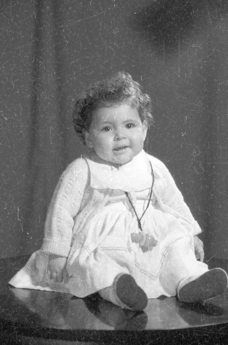 Retrato de estudio de Plano entero de un bebé con Vestido posando sentada sobre una mesa. Tiene su chupete colgando en el cuello