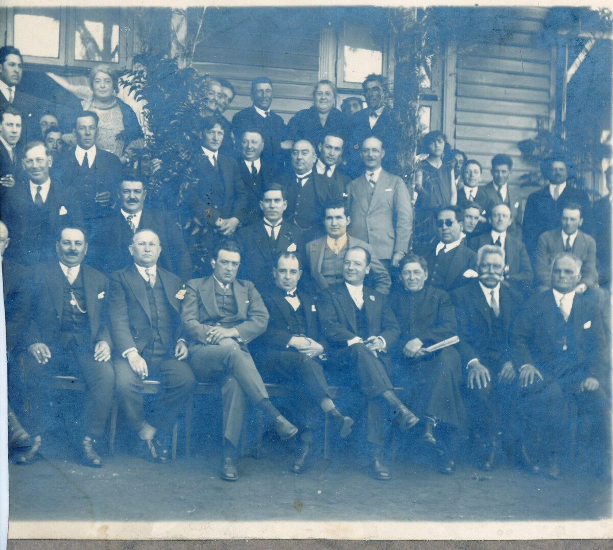 Retrato grupal de plano entero de Hombres y Mujeres posando en las afueras de una casa de madera no identificada.