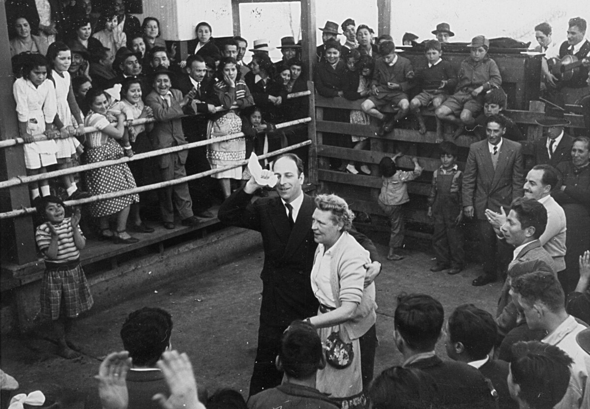 plano general de Olga Boettcher y un hombre vestido de Traje, realizando el paseo durante un baile de cueca. A su alrededor, un grupo numeroso de hombres, mujeres, Niñas y Niños aplauden como es costumbre para avivar el baile. Se encuentran en el interior de un recinto no identificado