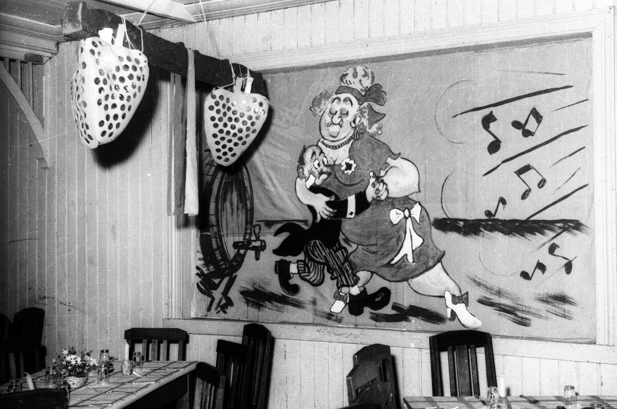 Plano entero de la pared de un recinto con un cuadro que muestra a una mujer grande bailando con un hombre pequeño, en el lugar hay mesas y sillas.