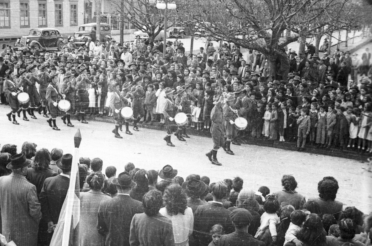 plano general de un grupo de Niños desfilando en hileras vestidos de scout mientras tocan sus instrumentos. A ambos lado de la calle por donde van desfilando hay un grupo número de Mujeres, Hombres, Niños y Niñas observándolos transitar. Todos ellos están ubicados en una de las calles laterales de la Plaza de La Concordia en La Unión