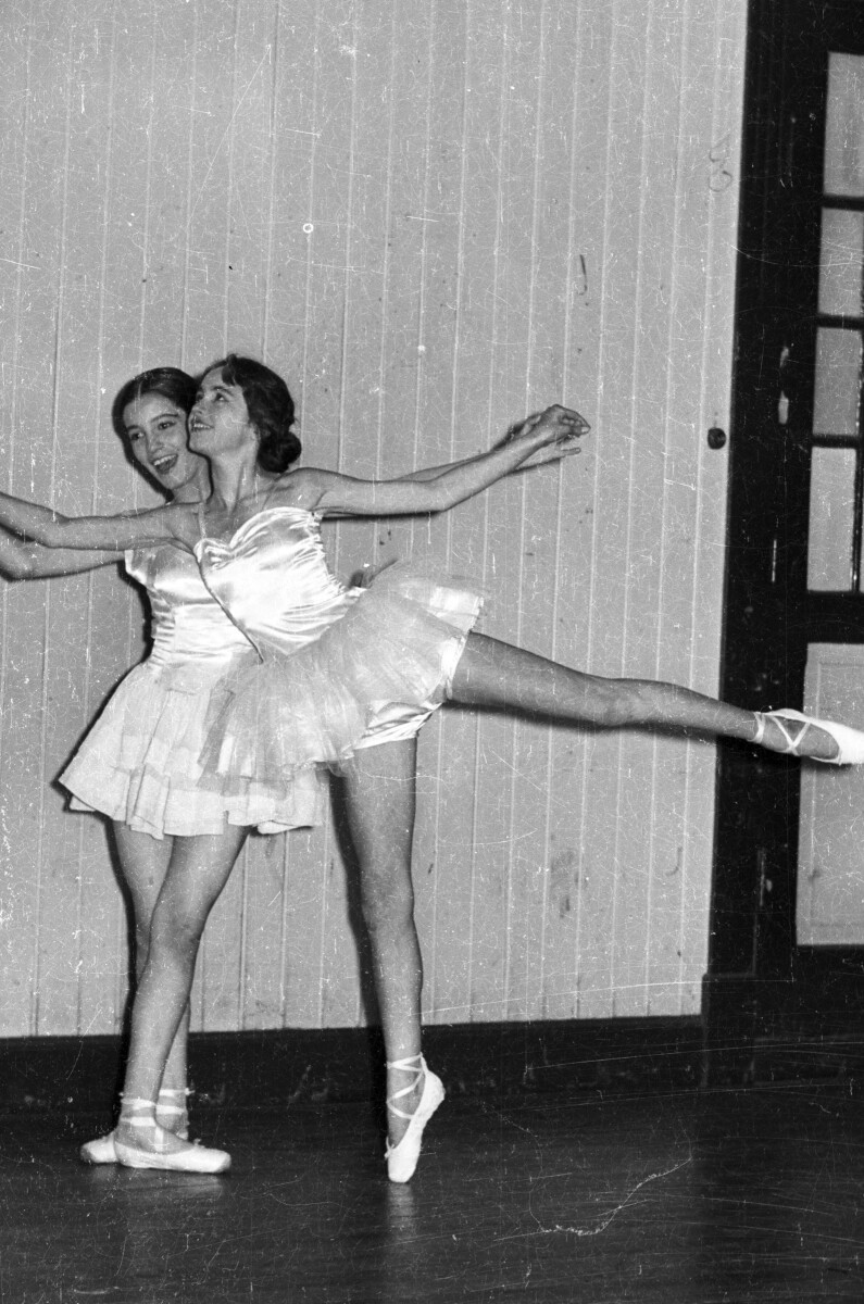 Plano entero de dos mujeres caracterizadas como bailarinas de ballet, paradas en posición de baile junto a una pared. Se encuentran en una estructura no identificada, durante una velada de presentaciones artísticas