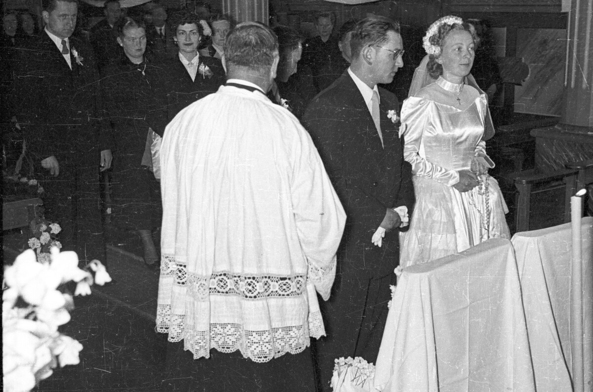 Plano entero de un acolito parado a un costado de una pareja de novios, quienes se encuentran parados junto a un reclinatorio en el altar. En el fondo un grupo numeroso de sus invitados se encuentran de pie junto a las bancas. Están en el interior de una parroquia no identificada, durante una misa de celebración de matrimonio