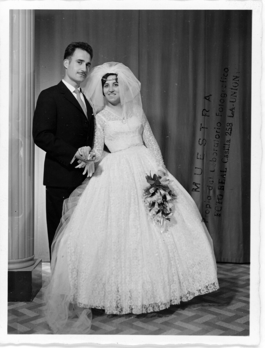 Retrato de estudio de Plano entero de una pareja de novios tomados de las manos. La novia lleva consigo un ramo de flores. Ambos sonríen y se encuentran parados junto a un pilar