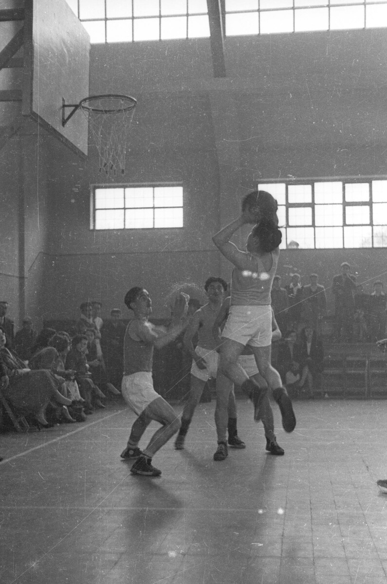 Plano entero de un hombre en el aire, lanzando el balón hacia el aro. Junto a él, hay tres hombres que lo rodean mientras realizan la jugada. En un costado de la cancha, hay un grupo de Hombres, Niños y Mujer sentados observando el partido de baloncesto el cual se está llevando a cabo en el interior del gimnasio de la Escuela Superior de Niñas Nº2