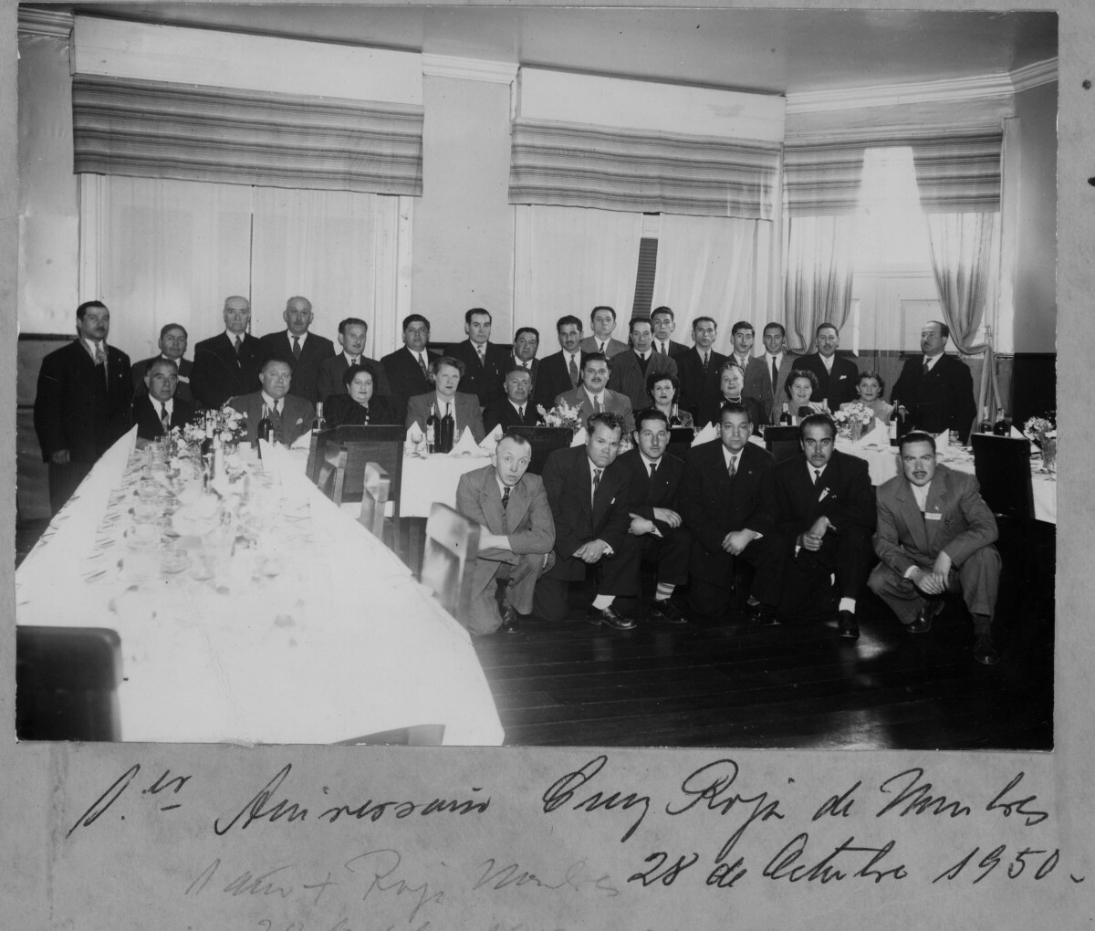 Retrato grupal de plano entero de un grupo de hombres pertenecientes a la cruz roja, posando en compañía de algunos mujeres, alrededor y en el frente de una larga mesa. La segunda mujer sentada de izquierda a derecha es Olga Boettcher, quien también es voluntaria de la institución. Se encuentran en el interior de una infraestructura durante la celebración del primer aniversario de los miembros masculinos.