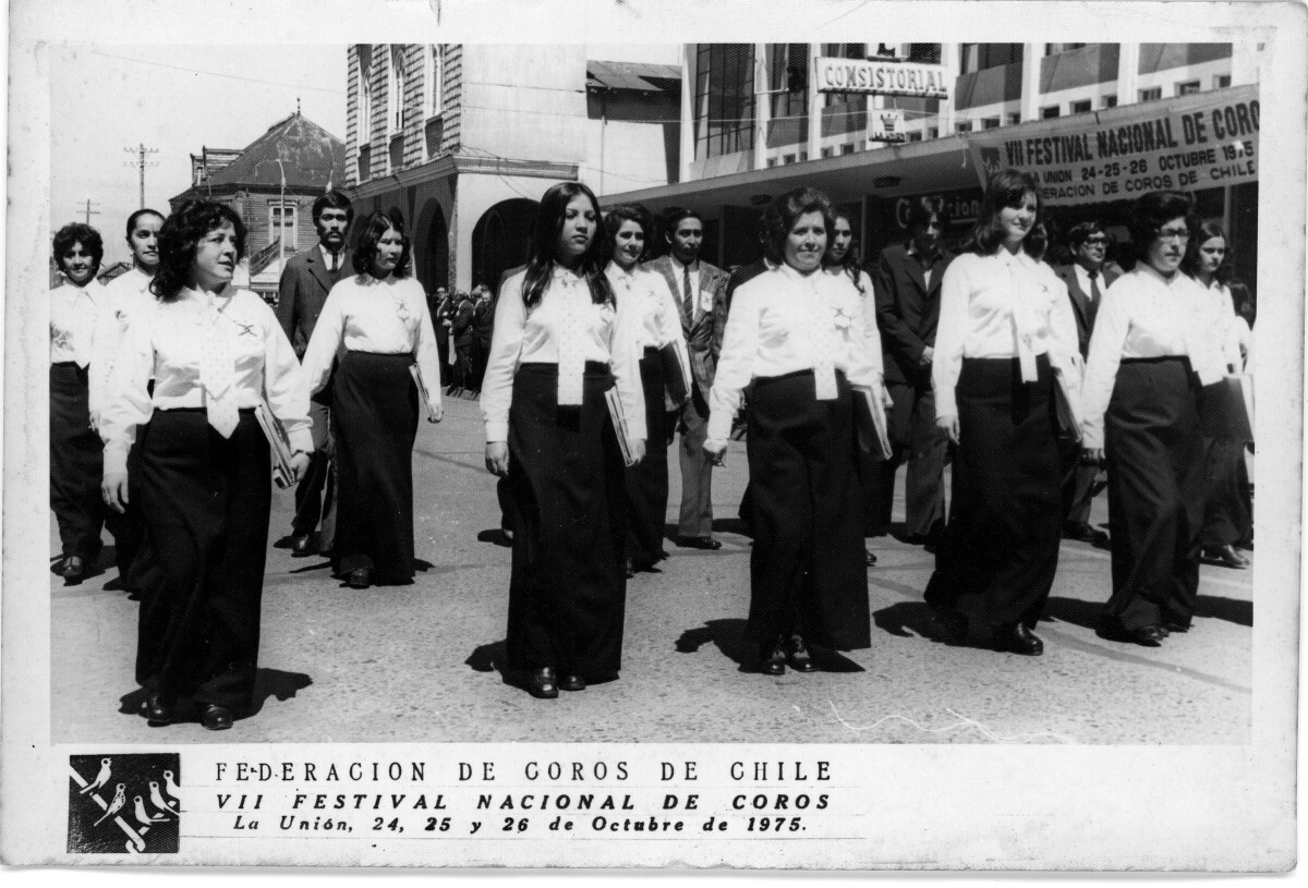 Plano general de integrantes de un coro, desfilando por calle Prat. En el fondo, se ve un lienzo colgado en el frontis de unas tiendas comerciales, el cual dice "VII festival de coros La Unión, 14 15 y 26 de Octubre de 1975, Federación de coros de Chile".