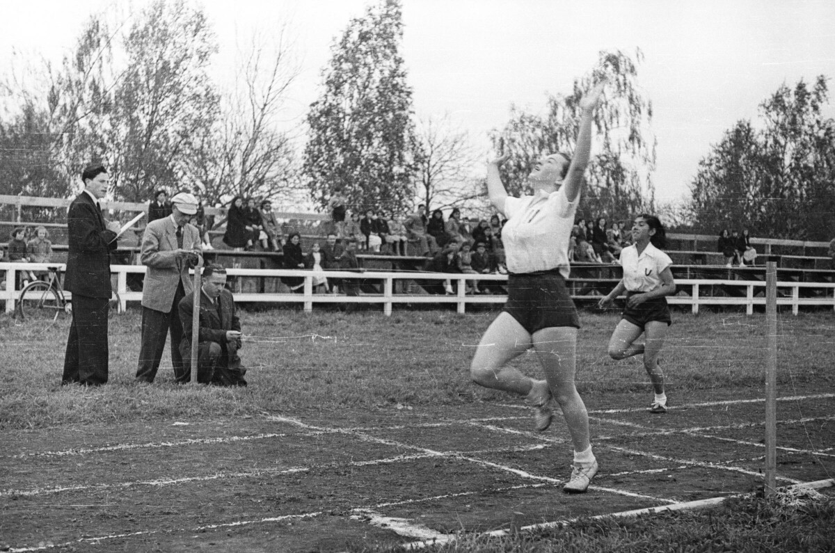 plano general de una mujer levantando las manos tras haber llegado a la línea de meta de la carrera, tras ella se aproxima otra mujer corriendo. En un costado hay tres hombres junto a la pista, dos de ellos anotan, mientras que el tercero se encuentra en cunclillas fumando un cigarro mientras observa, las graderías del fondo están ocupadas por un grupo de Mujeres, Hombres, Niños y Niñas que observan durante la jornada de competiciones de juegos olímpicos
