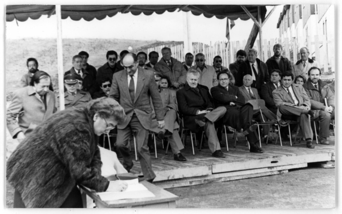plano general de Elfrida Ojeda sobre una mesa mientras firma un libro. En el fondo, hay un grupo de Hombres, Mujeres y dos oficiales de carabineros sentados sobre una tarima. Se encuentran en el interior de un recinto no identificado durante la entrega de unas viviendas sociales