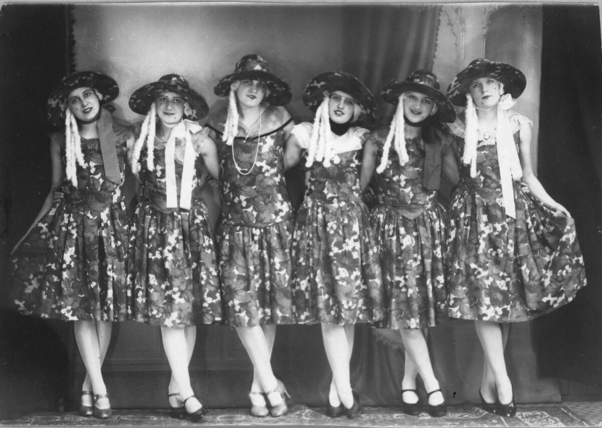 Retrato de plano entero de un grupo de mujeres posando abrazadas. Todas visten de igual forma, con un sombrero y un vestido floreado. En cada extremo, ambas mujeres levantan la punta de sus faldas con una mano, Se encuentran posando paradas sobre una alfombra