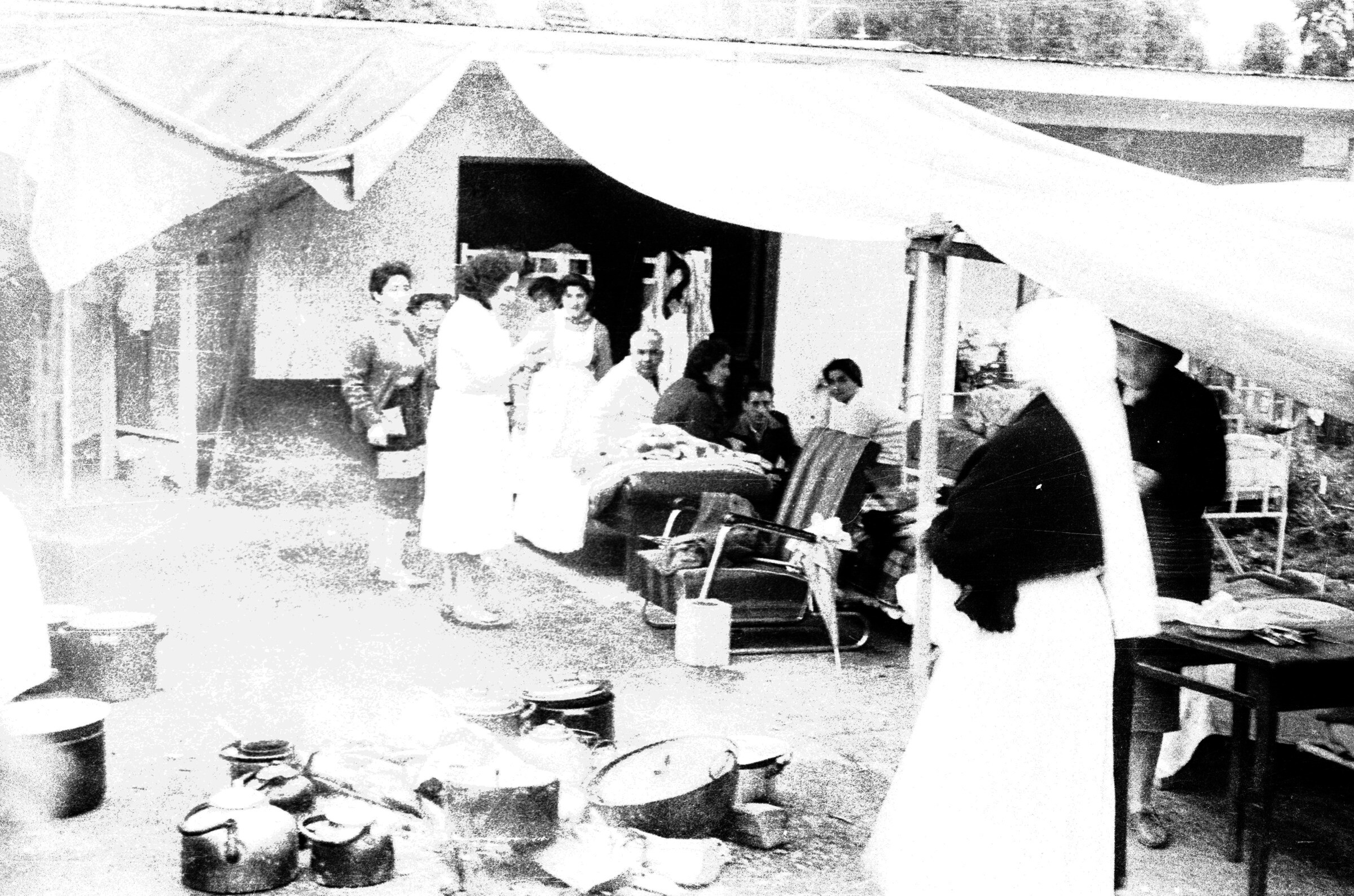 Plano general de hombres y mujeres sentadas y de pie en las afueras de hospital improvisado. En el centro hay ollas y teteras alrededor de una fogata.