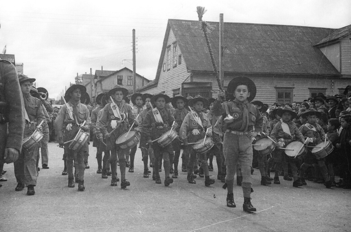 Plano entero de un Niño guiando con una guaripola a la orquesta sinfónica de la brigada de scout infantil, quienes van avanzando detrás de él. En un costado hay un grupo de Niñas, Niños, Mujer y Hombres observándolos mientras desfilan
