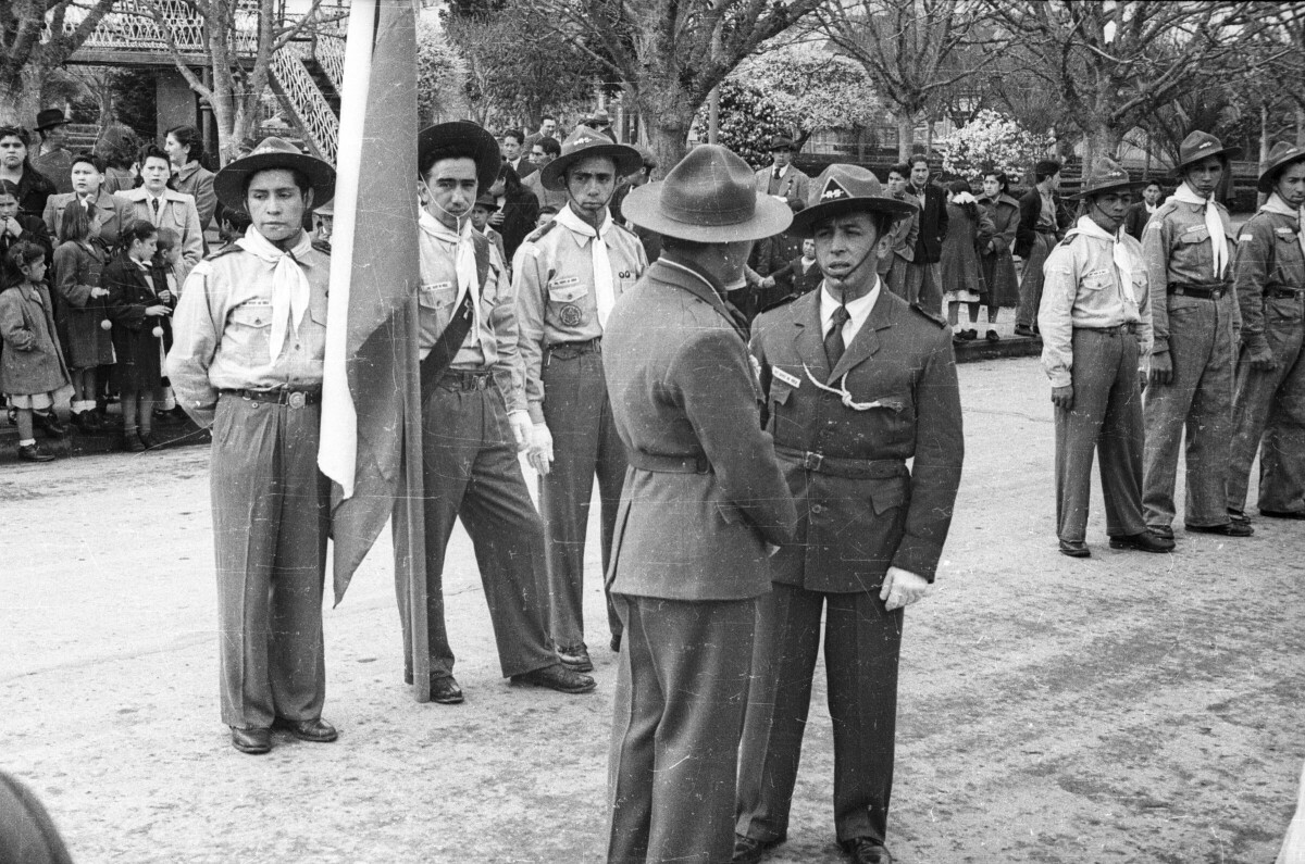 Plano entero de dos hombres pertenecientes a la brigada de scout conversando de pie, detrás de ellos hay otro grupo de hombres parados en filas llevando puesto su uniforme de la brigada, uno de ellos, está sosteniendo el mástil de la bandera chilena. Junto a la calle donde están, sobre la vereda, hay un grupo de Niñas, Niños , Mujeres y Hombres observándolos desde La Plaza de La Concordia de La Unión