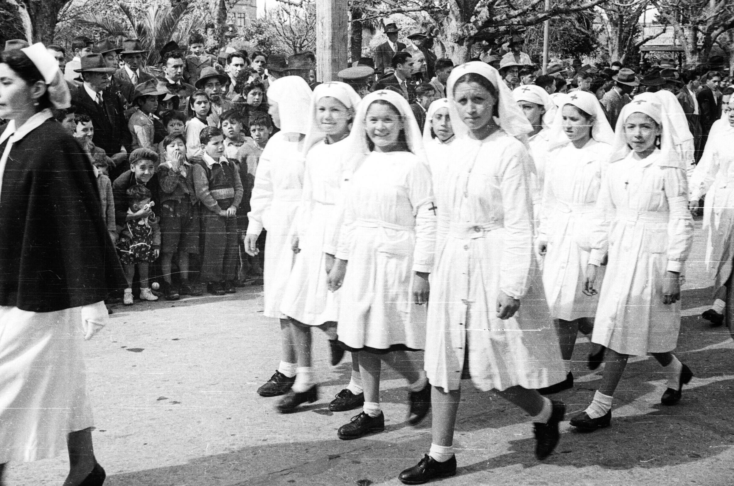 plano general de un grupo de Niñas caminando vestidas de blanco con una túnica en su cabeza que tiene dibujada una cruz sobre la frente. Ellas pertenecen a la Cruz Roja Juvenil de la Escuela Nº2. En un costado de la calle hay un grupo de Niños, Niñas, Hombres y Mujeres observándolas de pie sobre la vereda durante un desfile por las calles de La Unión