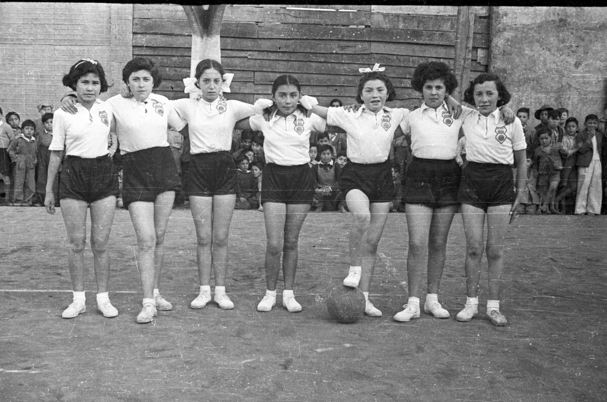 Retrato de plano entero de un equipo de baloncesto de Niñas, están posando todas de pie abrazadas, una de ellas tiene su pie sobre la pelota. En el fondo hay un grupo de Niños apoyados en una pared de la iglesia, hay un grupo de ellos que están descalzos mientras observan en un costado de la cancha