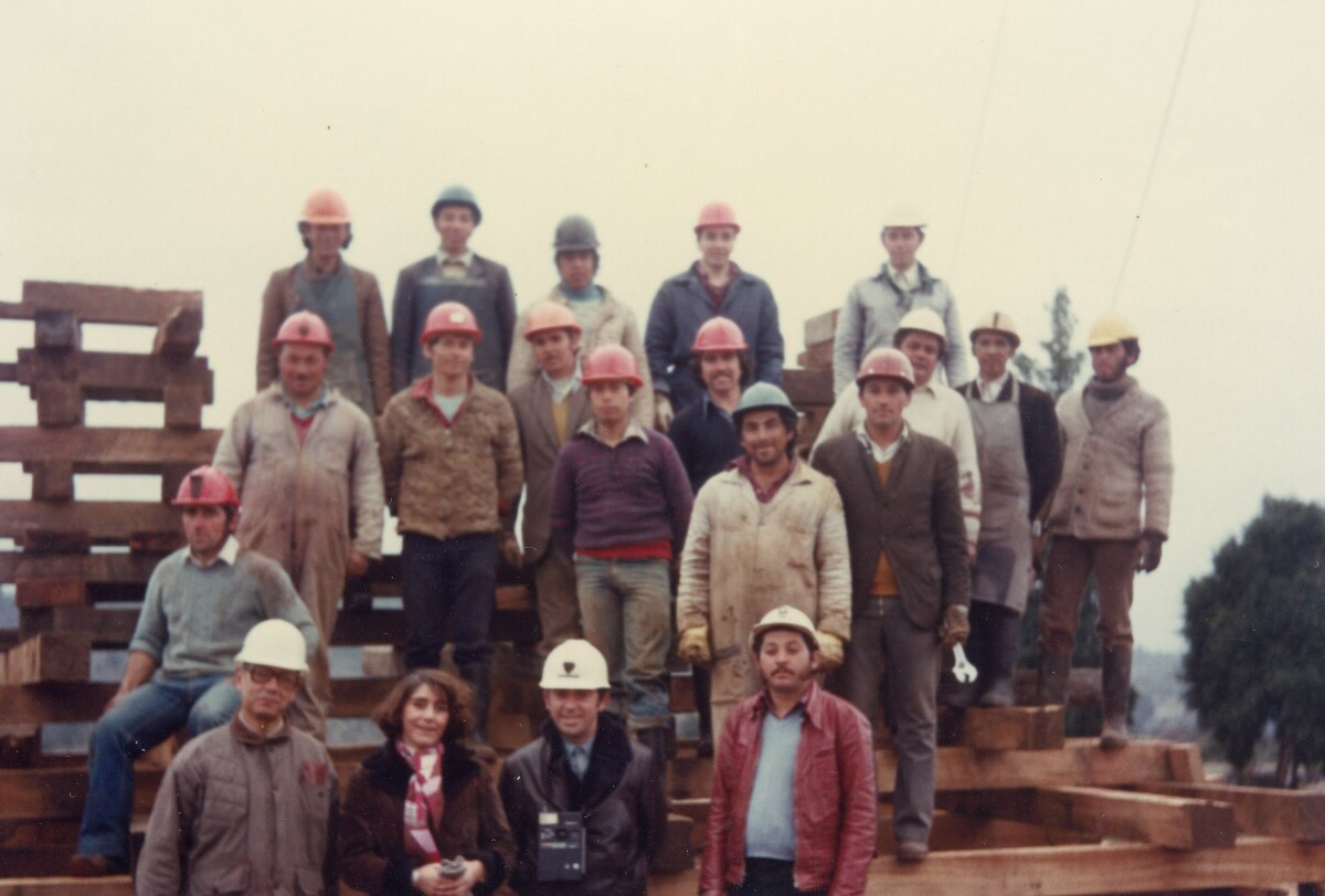 plano general de un grupo numeroso de mineros posando junto a una mujer y dos hombres de casco blanco, sobre una estructura de madera ubicada en las dependencias de la Compañía carbonífera San Pedro de Catamutún