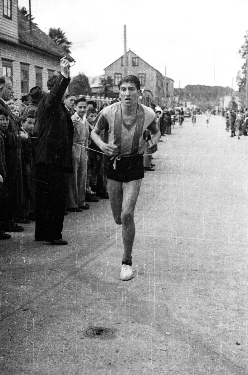 Retrato de plano entero de un hombre llegando a la línea de meta, junto a él hay un grupo de personas compuesto por Hombres, Mujeres, Niños y Niñas observando la carrera a lo largo de la calle donde se está realizando la competencia.