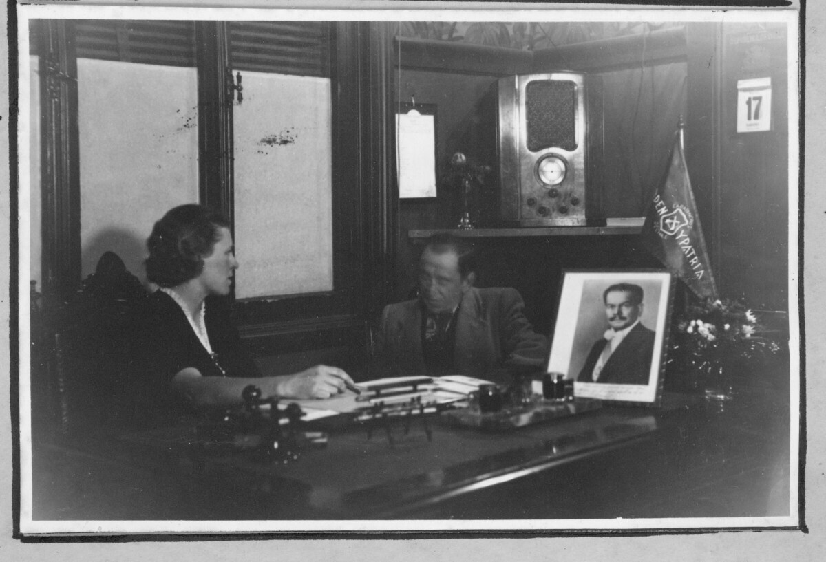 Plano entero de Olga Boettcher, conversando con un hombre. Ambos están sentados junto a un escritorio, sobre el cual hay documentos, un retrato de Pedro Aguirre Cerda y también hay un banderín de carabineros que dice -Orden y Patria-. En el fondo, hay una radio sobre la repisa de la esquina. Se encuentran en una oficina de una infraestructura no identificada