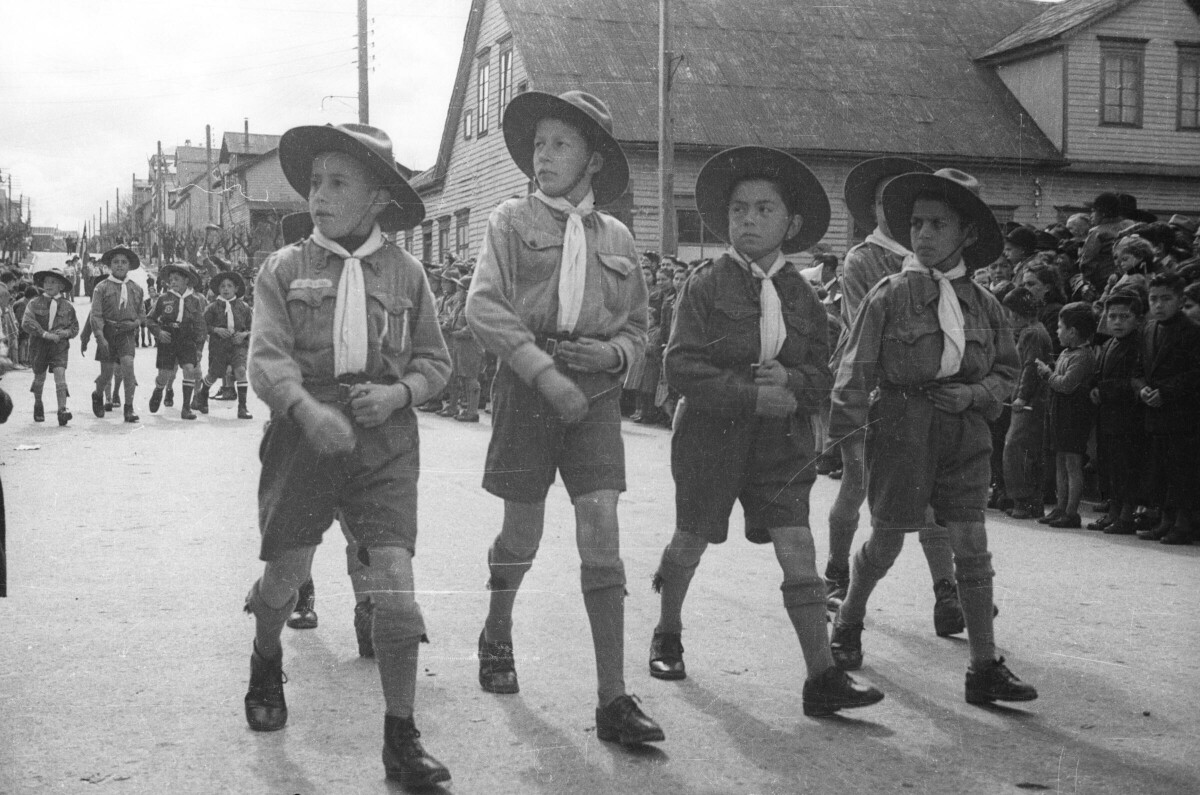 Plano entero de un grupo de Niños pertenecientes a la brigada de scout, caminando formados en dos hileras mientras miran hacia su costado derecho. Tras ellos, otro grupo de Niños va avanzando mientras son observados por un grupo numeroso de personas y un carabinero, quienes están ubicados a un costado de la calle por donde se está realizando el desfile.