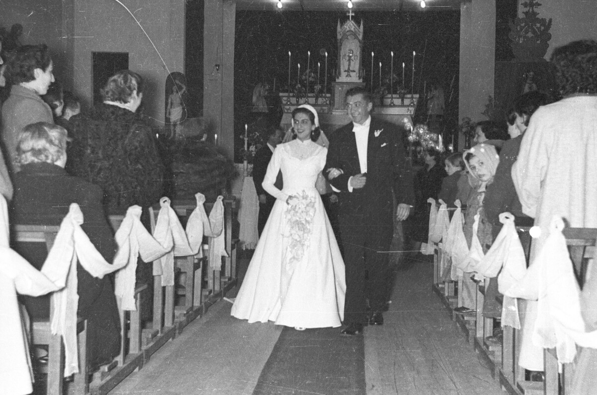 Plano entero de una pareja de recién casados. Van tomados del brazo caminando por la nave central mientras salen de la iglesia una vez finalizada la misa del sacramento.