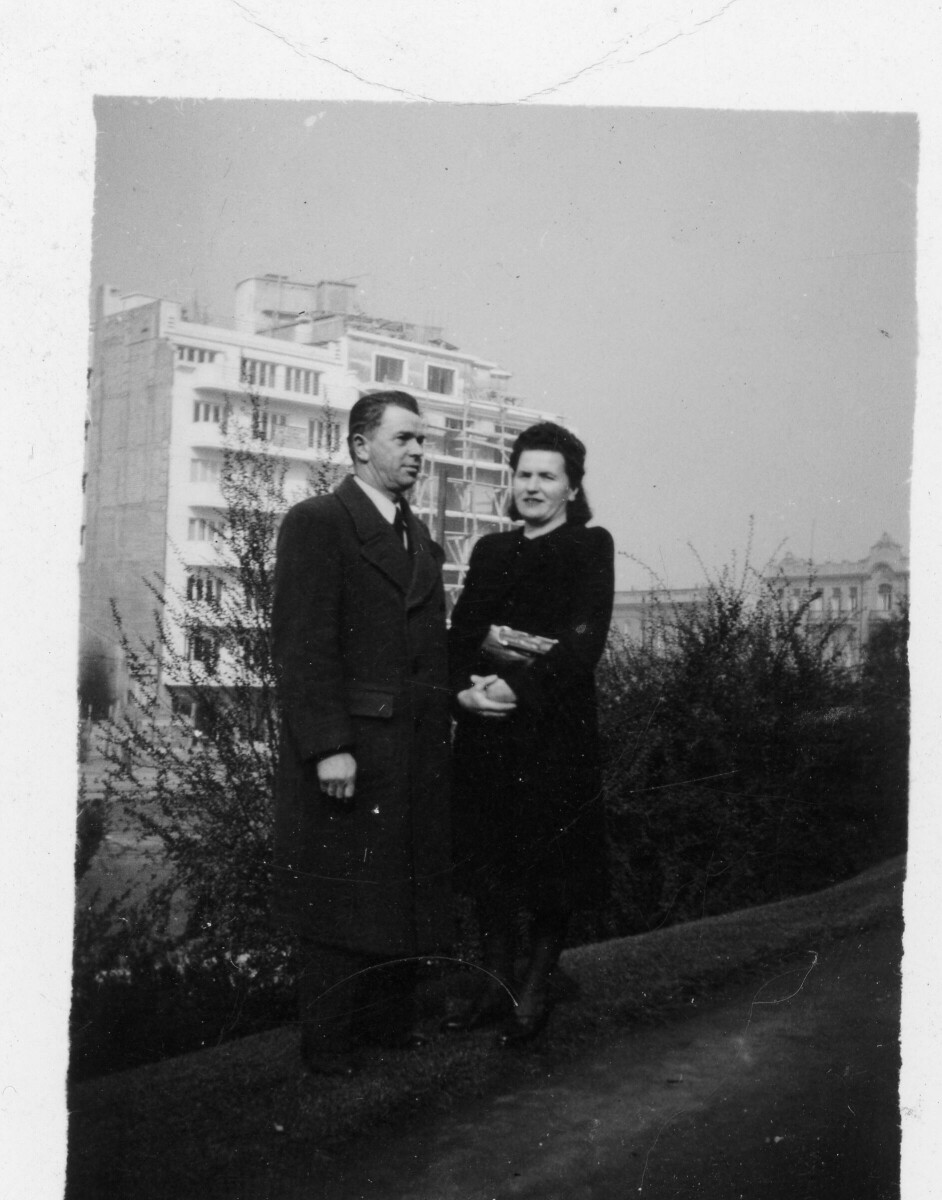 Retrato de plano entero de un hombre y una mujer posando al costado de un camino de tierra. A sus espaldas hay un edificio. Ciudad no identificada.