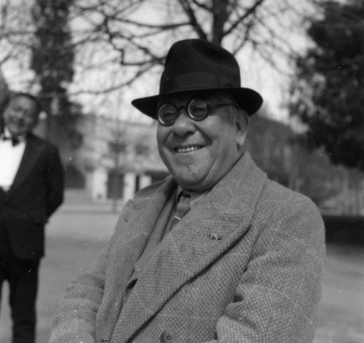 Retrato de plano medio largo  de un hombre sonriendo a la cámara. Lleva puesto un sombrero y lentes con aumento. En el fondo, hay un hombre de Traje sonriendo. Se encuentran ubicados a un costado de la plaza de La Concordia