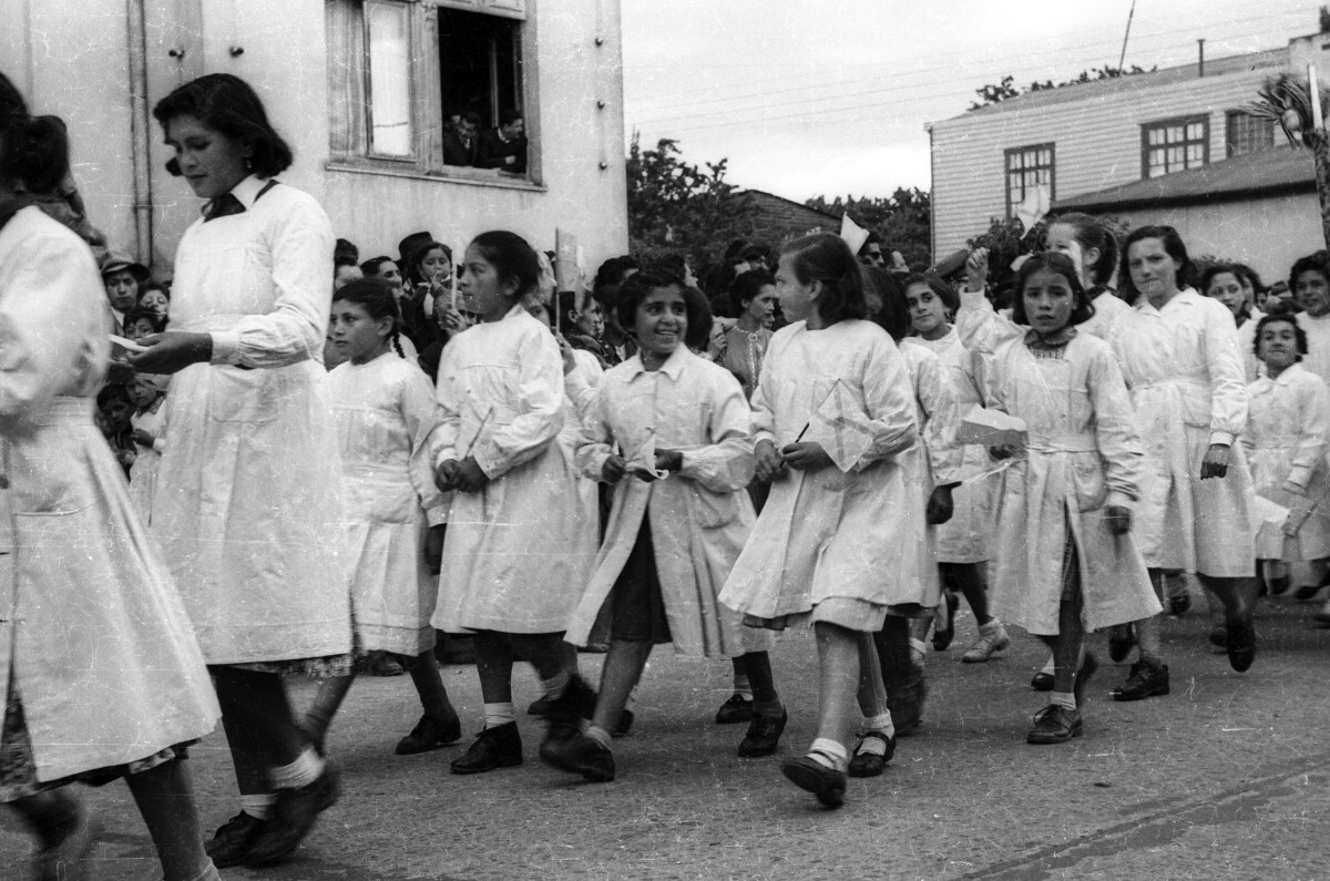 Plano entero de un grupo de niñas con delantal, desfilando formadas en hileras, con una bandera en las manos, por el frontis de la municipalidad
