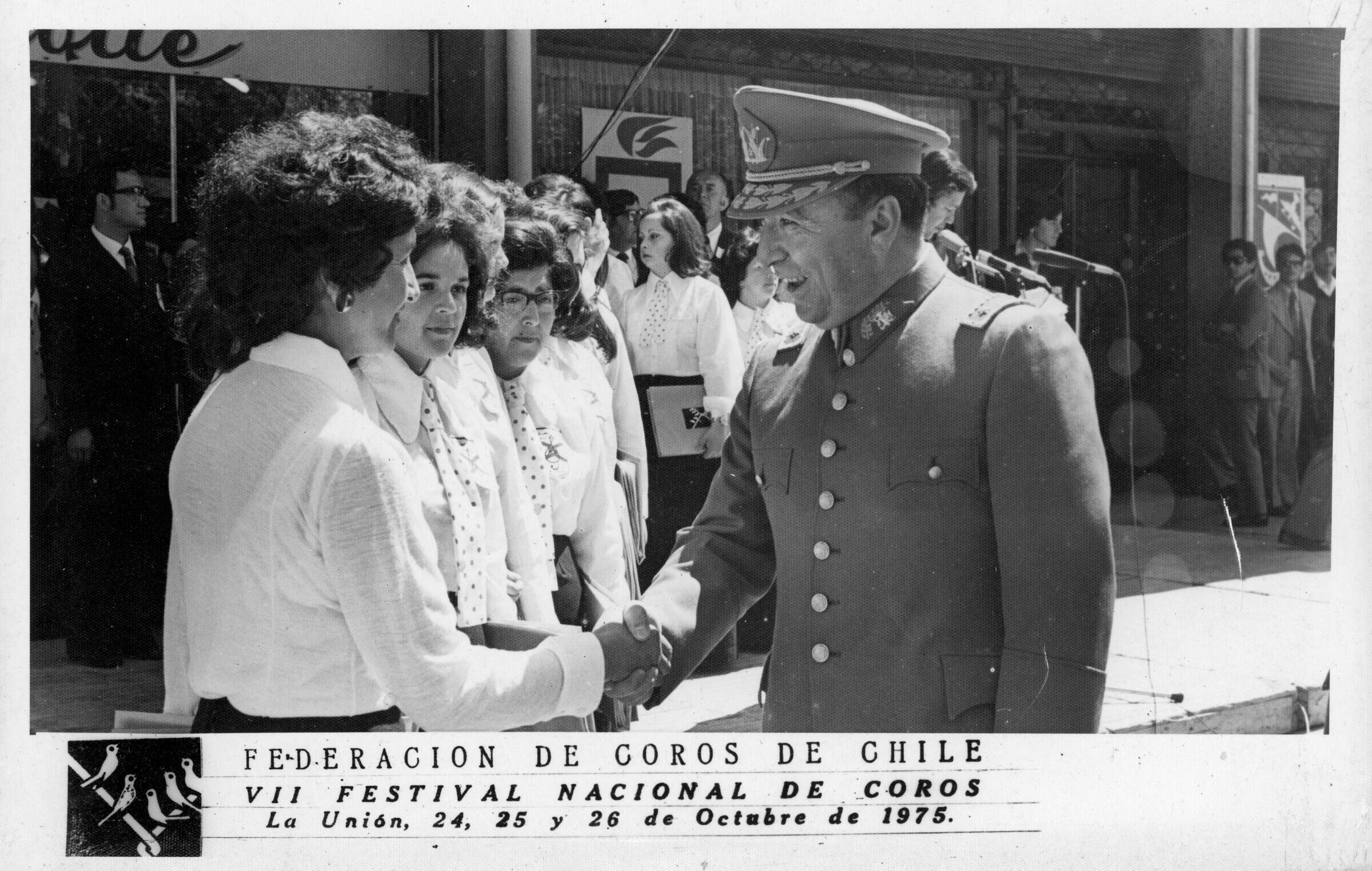 Plano entero de un uniformado de carabinero, estrechando la mano de una mujer perteneciente a un grupo coral no identificado. Junto a ella, se encuentran paradas otras mujeres, miembros del coro. Están ubicados en el frontis de unas tiendas comerciales, durante el VII festival nacional de coros realizado en La Unión