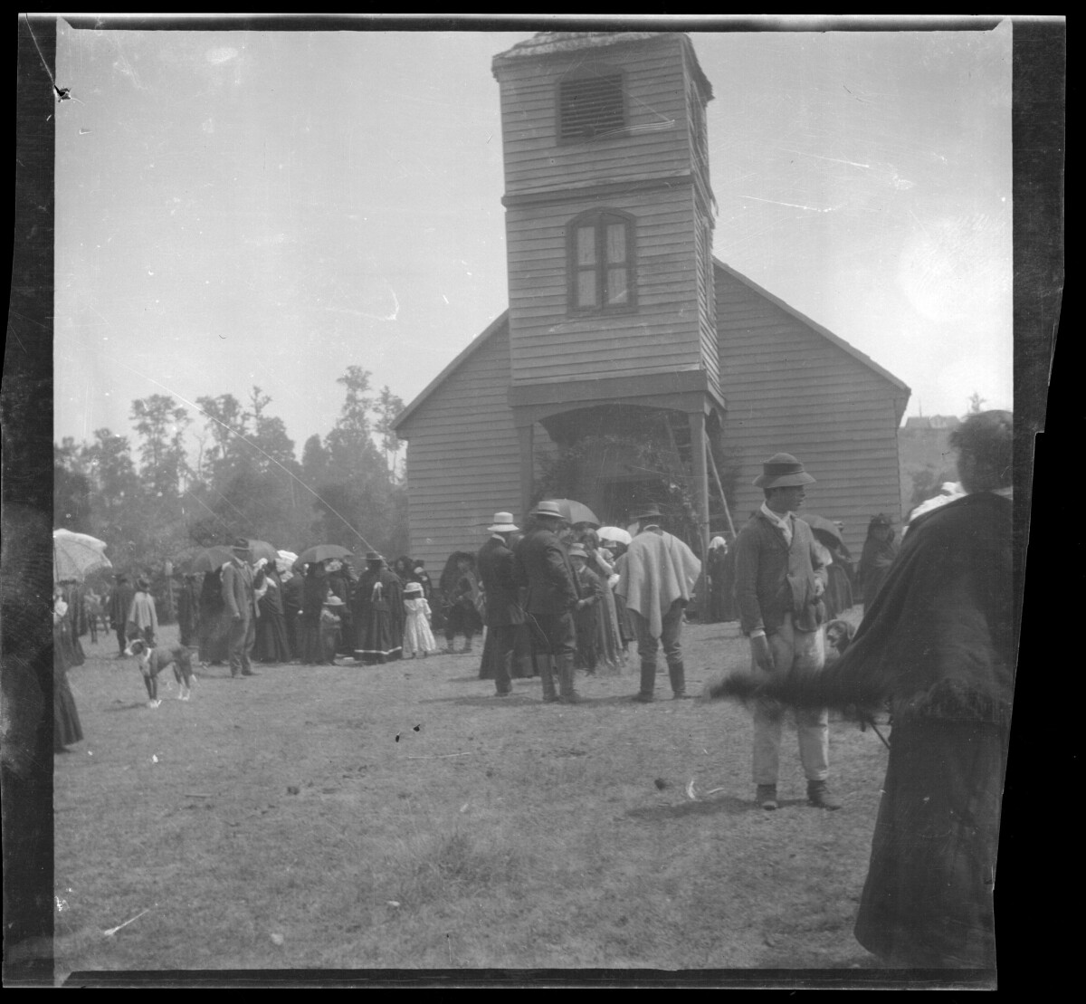 Multitud de personas en ceremonia religiosa a las afueras de una iglesia. Las personas usan vestidos, ponchos, sombreros y paraguas.