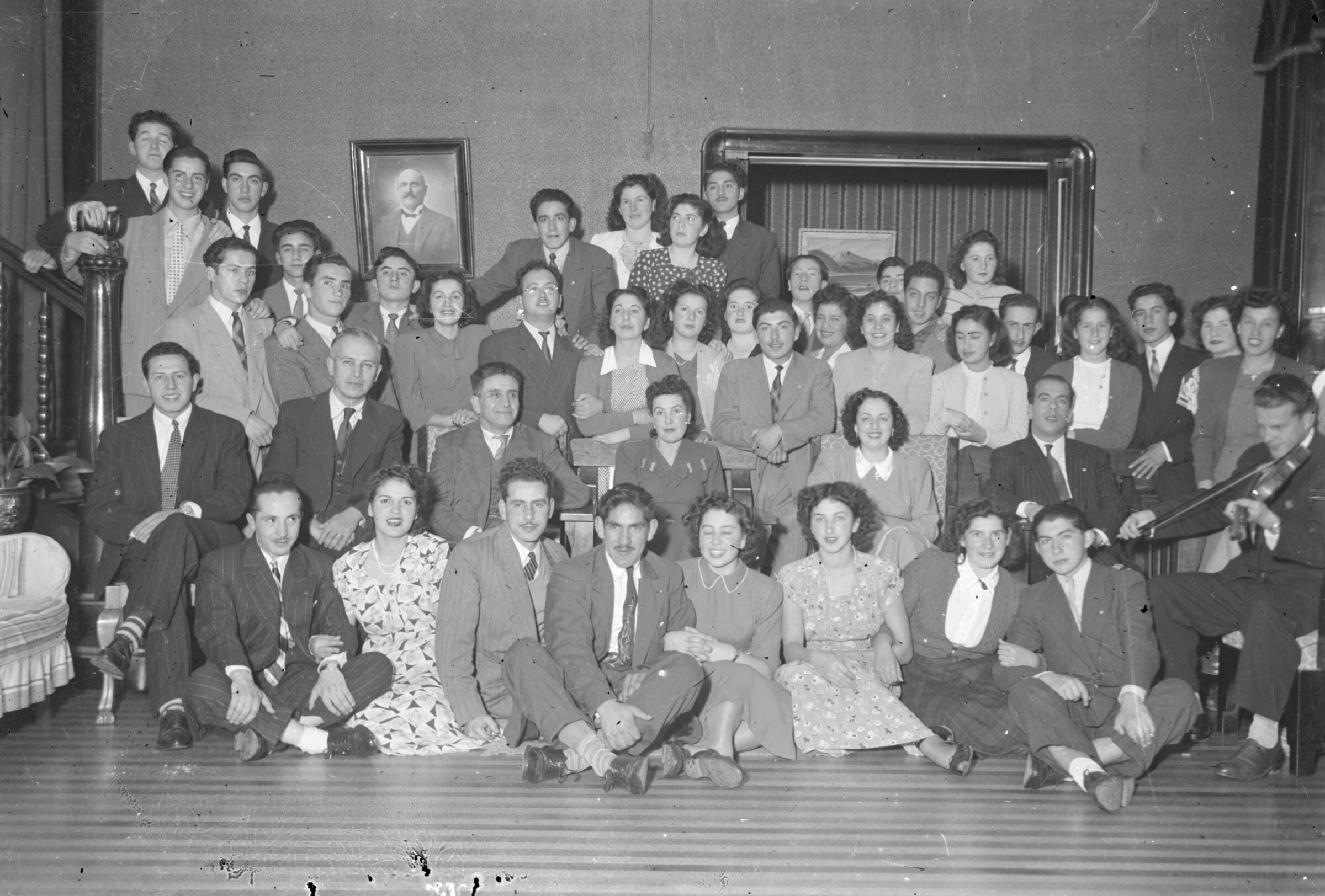 Plano general de un grupo de personas posando en la habitación de una casa. Uno de ellos toca un violín