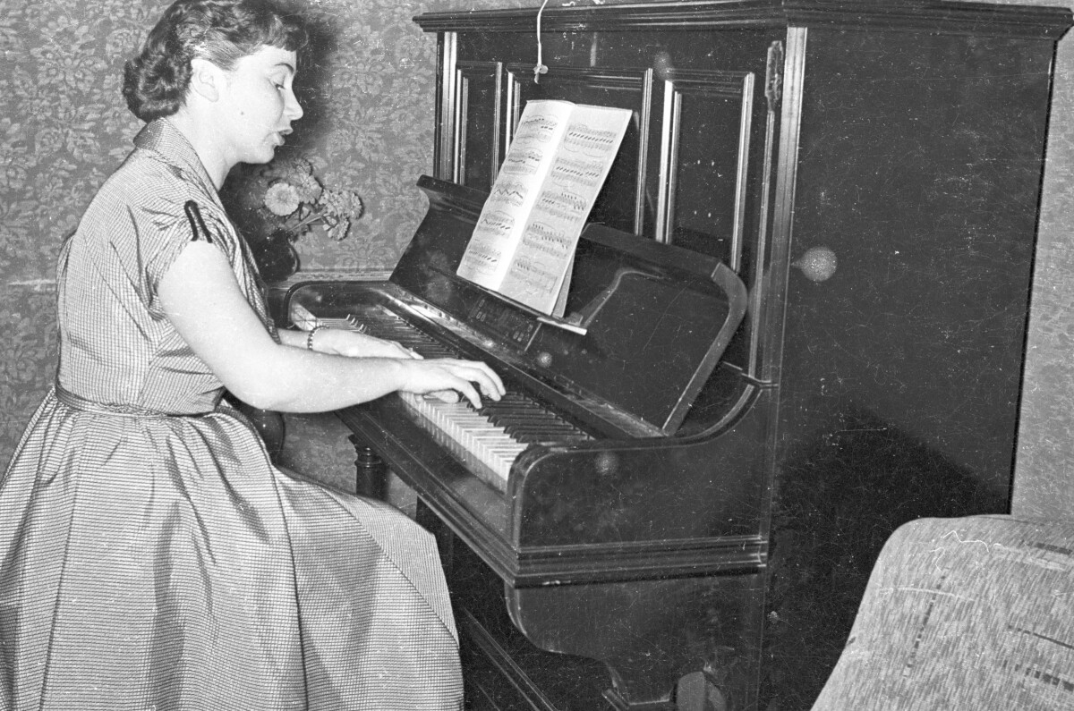 Plano medio largo de una mujer con Vestido sentada, tocando un piano de pared mientras canta. Frente a ella, se encuentra abierta una partitura. Está en el interior de una casa.