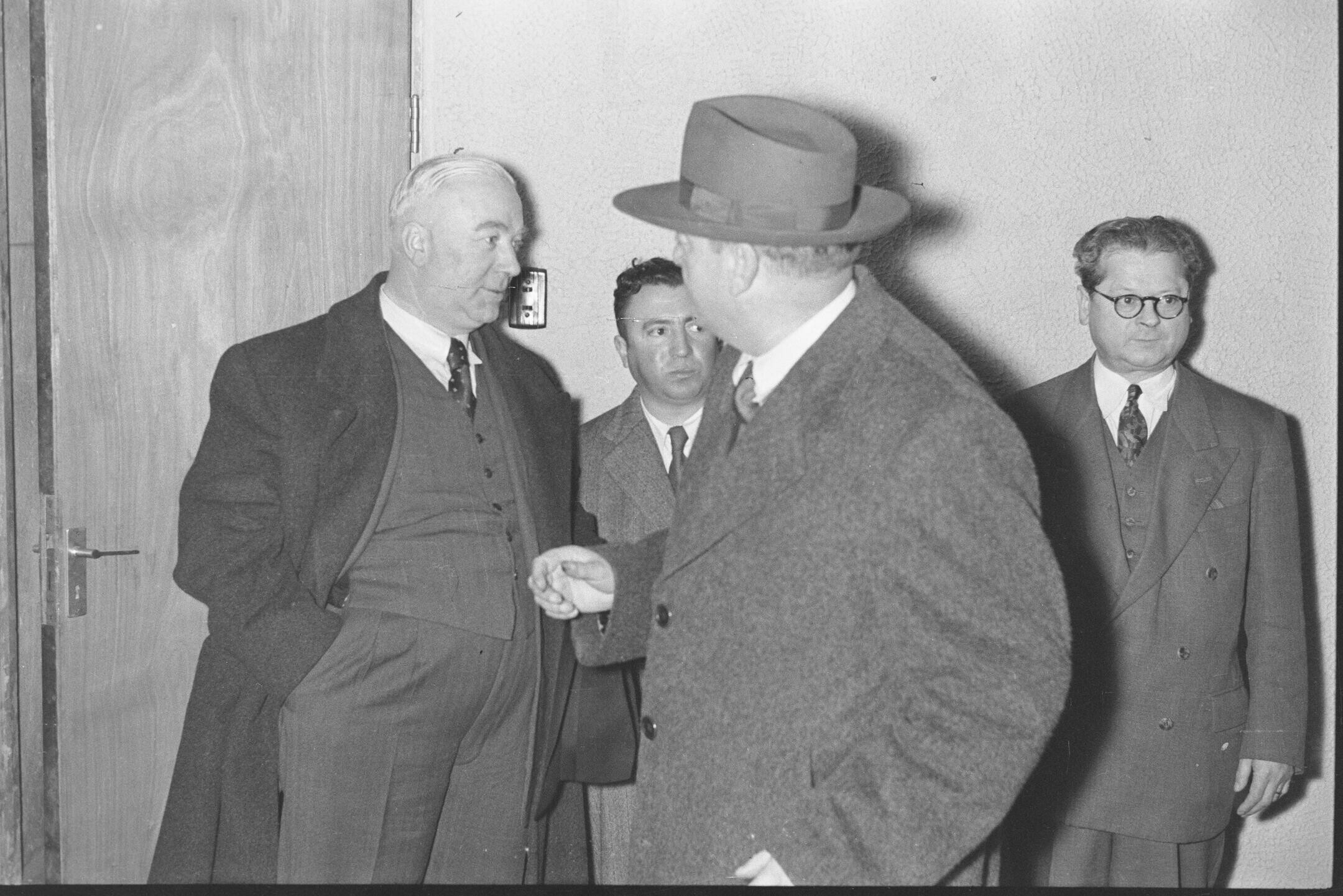 Plano entero de dos hombres conversando junto a otros dos hombres quienes se encuentran apoyados en un pared. Están en el interior del teatro O'Higgins