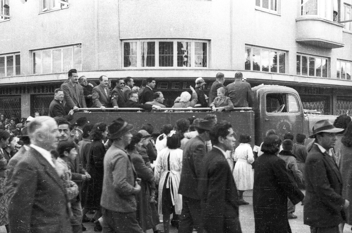 plano general de un grupo de personas y Carlos Vogel, en la parte trasera de un camión tres cuartos. Sobre la calle, una caravana los acompaña mientras avanzan por el frontis de la casa Masri
