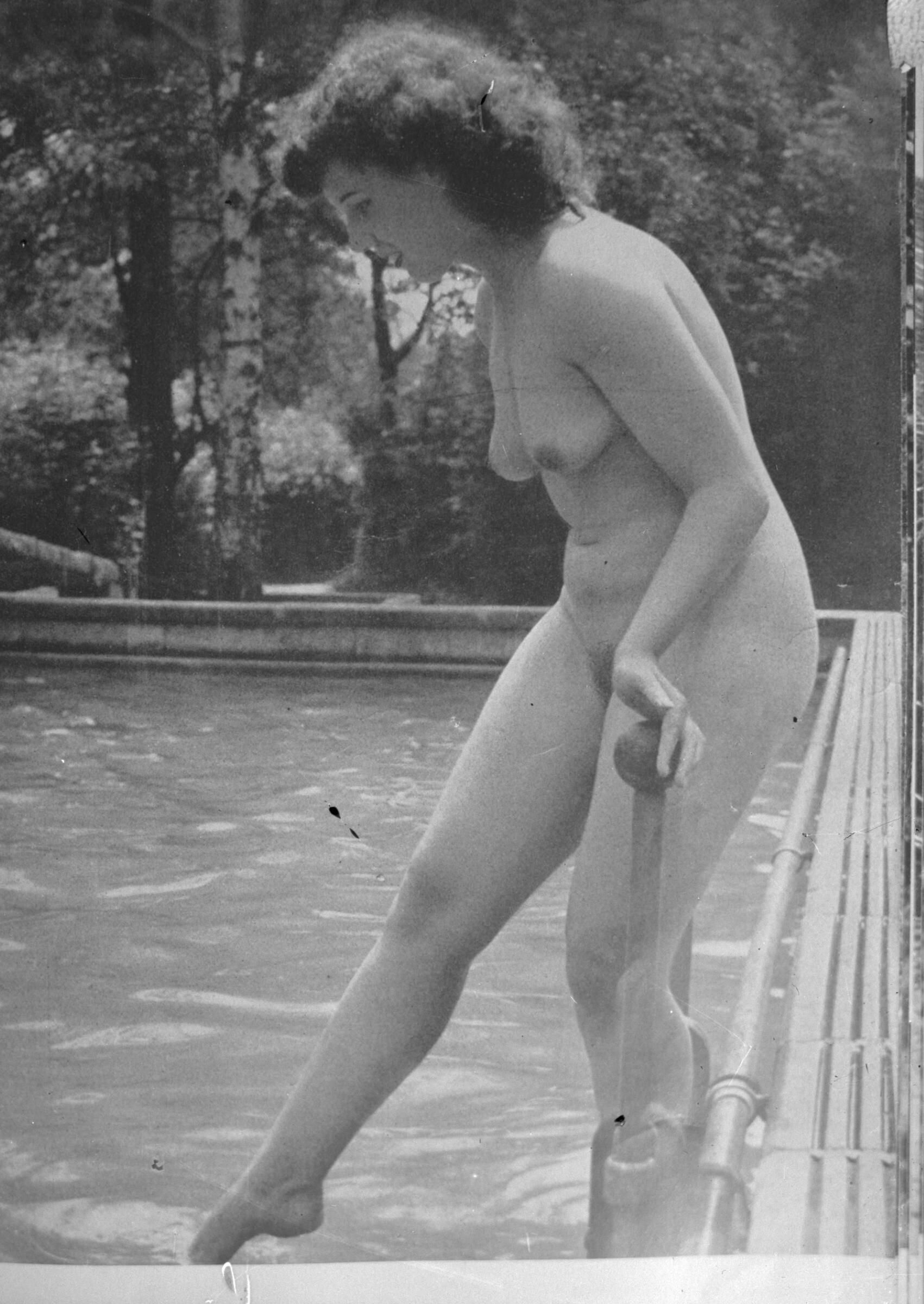 Reproducción de fotografía. Retrato de plano entero de una mujer desnuda entrando a una piscina