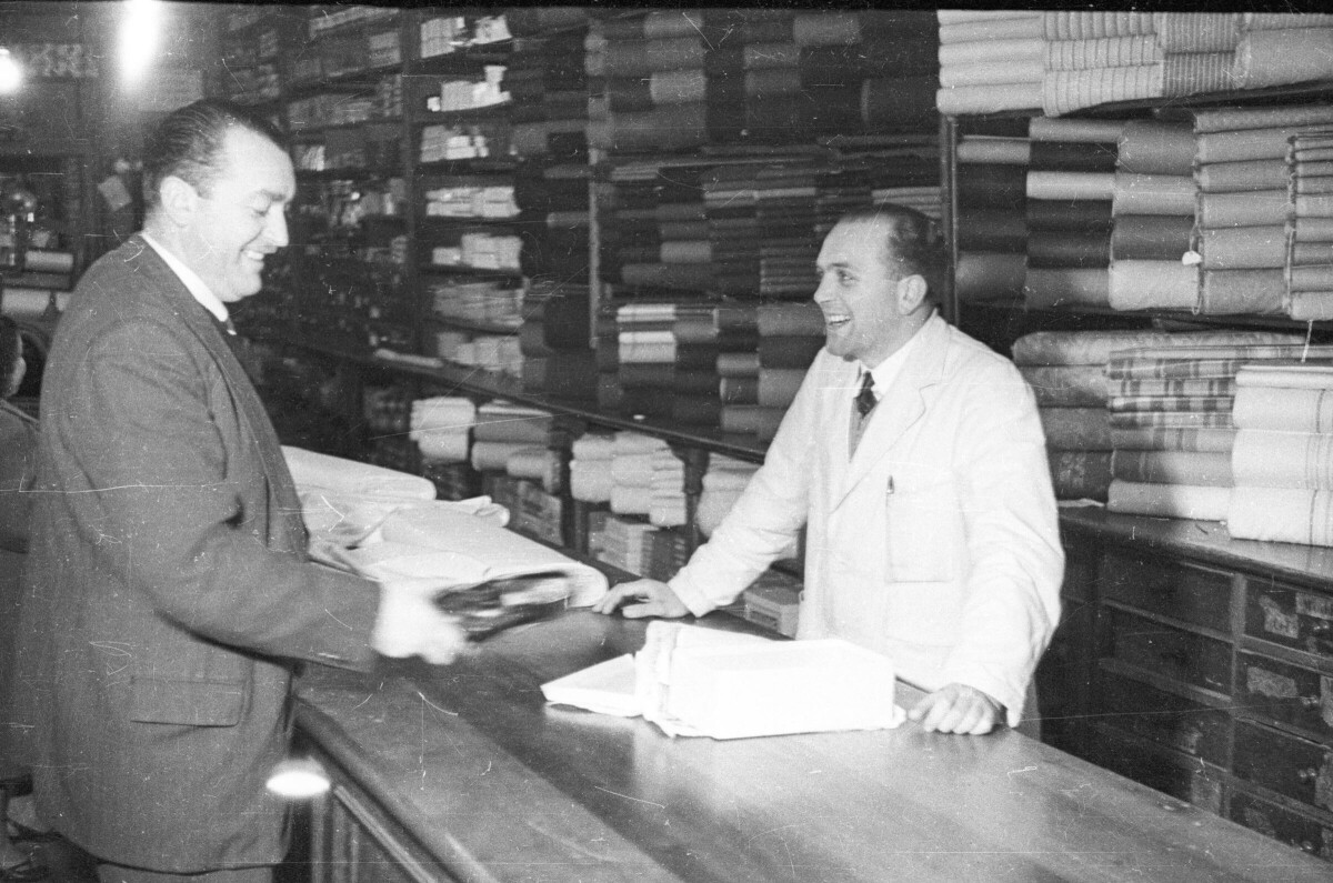 plano general de un hombre con un zapato en la mano, frente a el un vendedor lo atiende mientras ambos se ríen, en el fondo hay un estante con rollos de telas apilados en una tienda