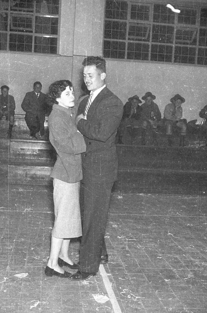 Plano entero de una mujer y un hombre bailando tomados de la mano. En las graderías del fondo hay un grupo de Niños y un hombre sentados. Están en el interior de un gimnasio.