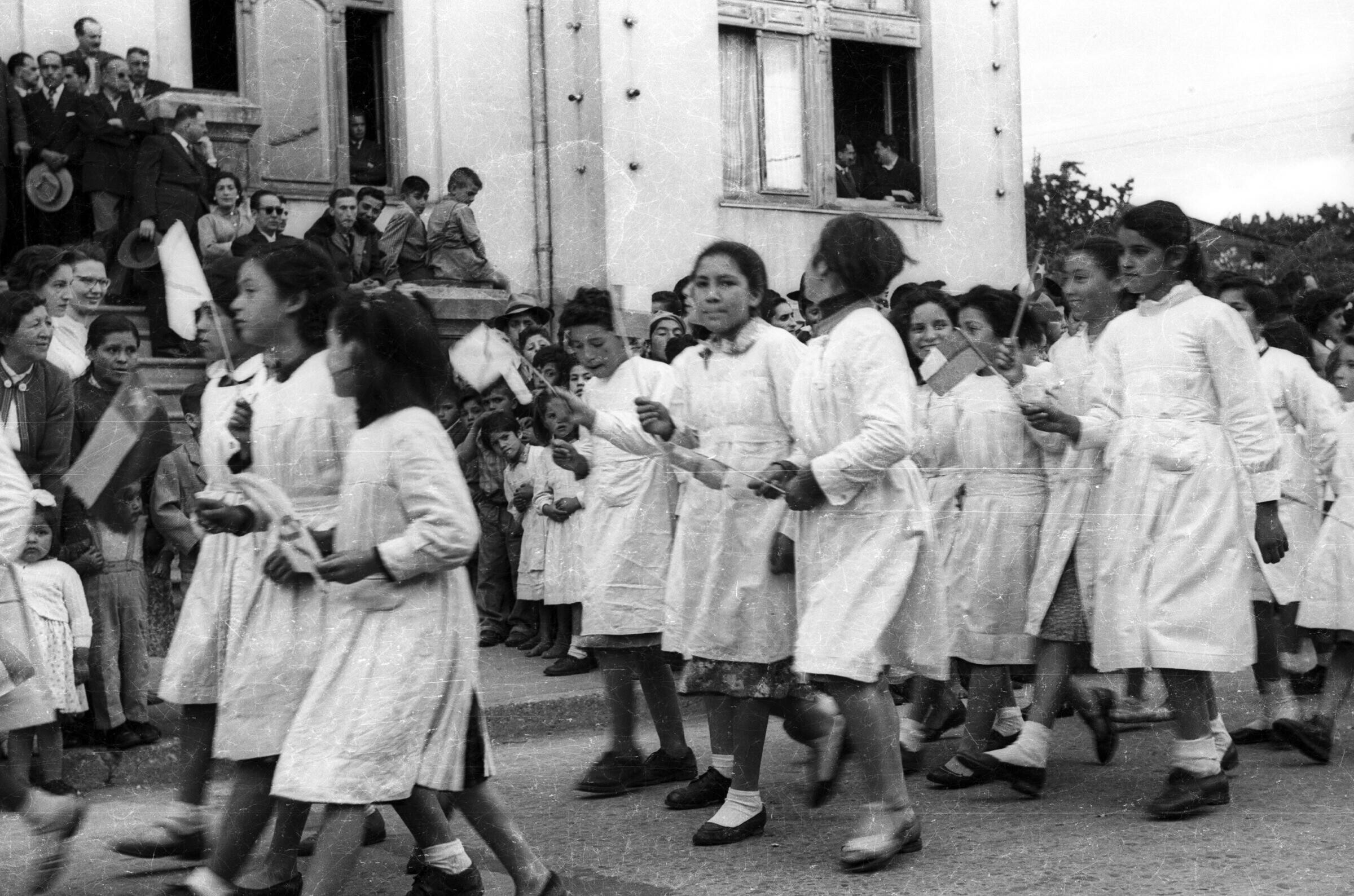 plano general de un grupo de niñas con delantal, desfilando formadas en hileras, con una bandera en las manos. En el fondo, un grupo de personas se encuentran observándolas en el frontis de la municipalidad