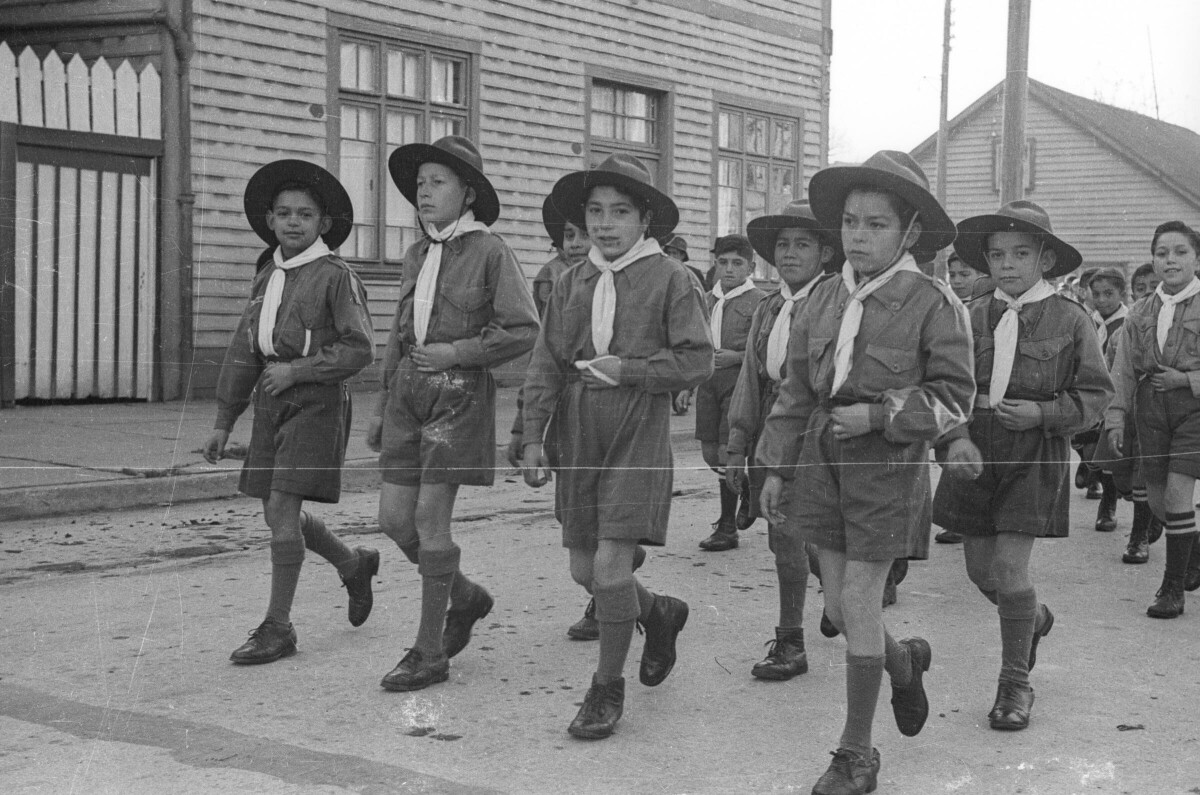Plano entero de un grupo de Niños pertenecientes a una brigada de scout desfilando en hileras por una calle pavimentada