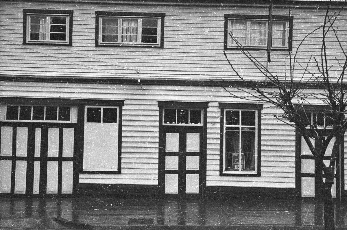 Vista general de una casa de dos pisos. En la ventana junto a la puerta hay un letrero con un mensaje ilegible y en el segundo piso hay una bandera izada. La fotografía fue tomada durante un día lluvioso