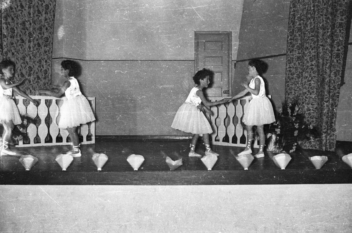 Plano entero de cuatro niñas bailando en pareja tomadas de la mano, están vestidas como bailarinas de ballet. Están bailando sobre un escenario en el interior de un colegio durante una actividad escolar