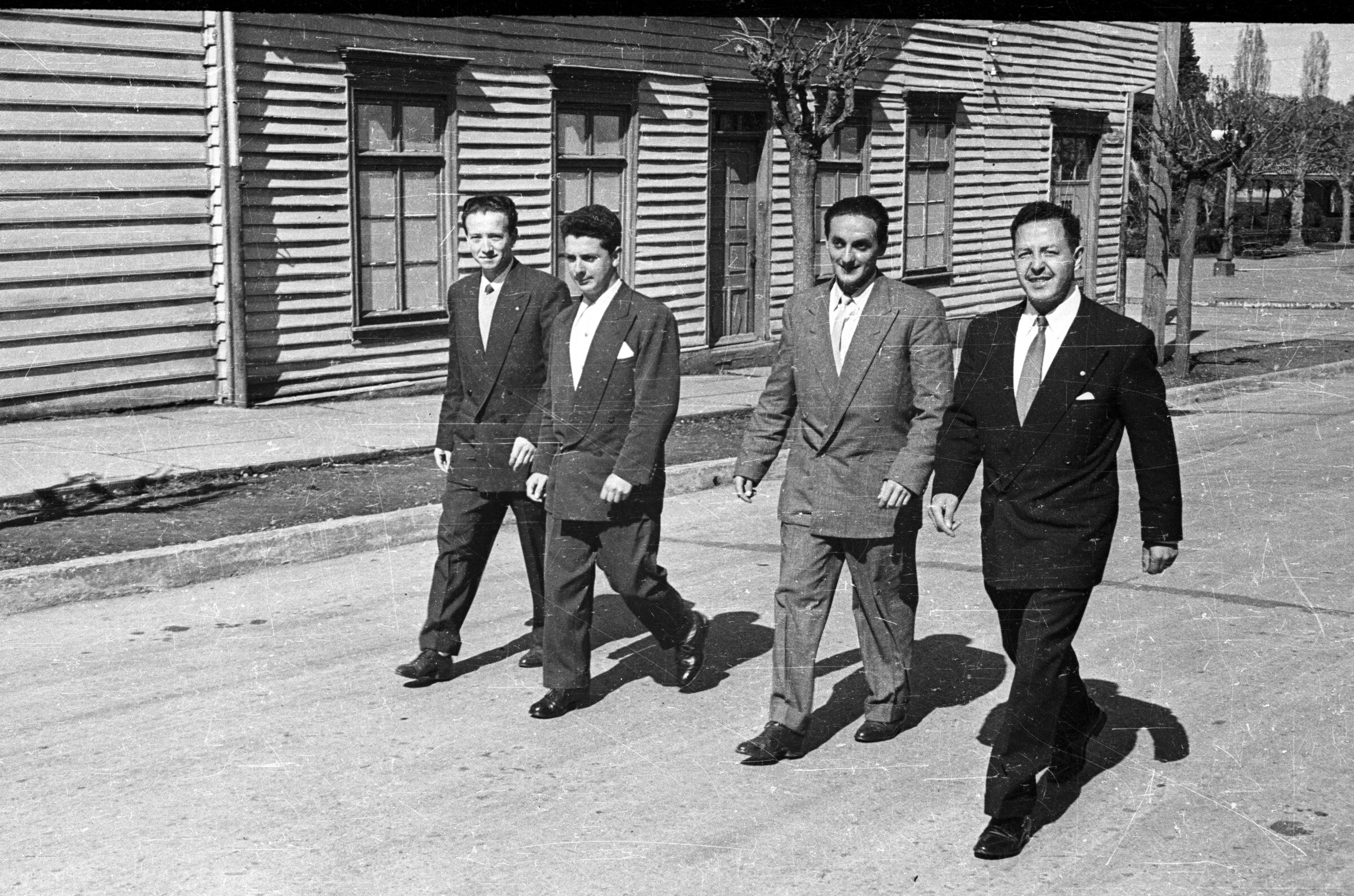 Plano entero de cuatro hombres caminando vestidos de traje en el medio de una calle pavimentada