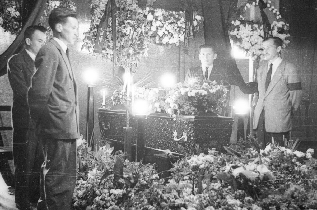 Plano entero de cuatro hombres v vestido de trajes, parados alrededor de un féretro. A su alrededor hay velas encendidas y coronas de flores adornando el lugar donde está llevando a cabo el velorio