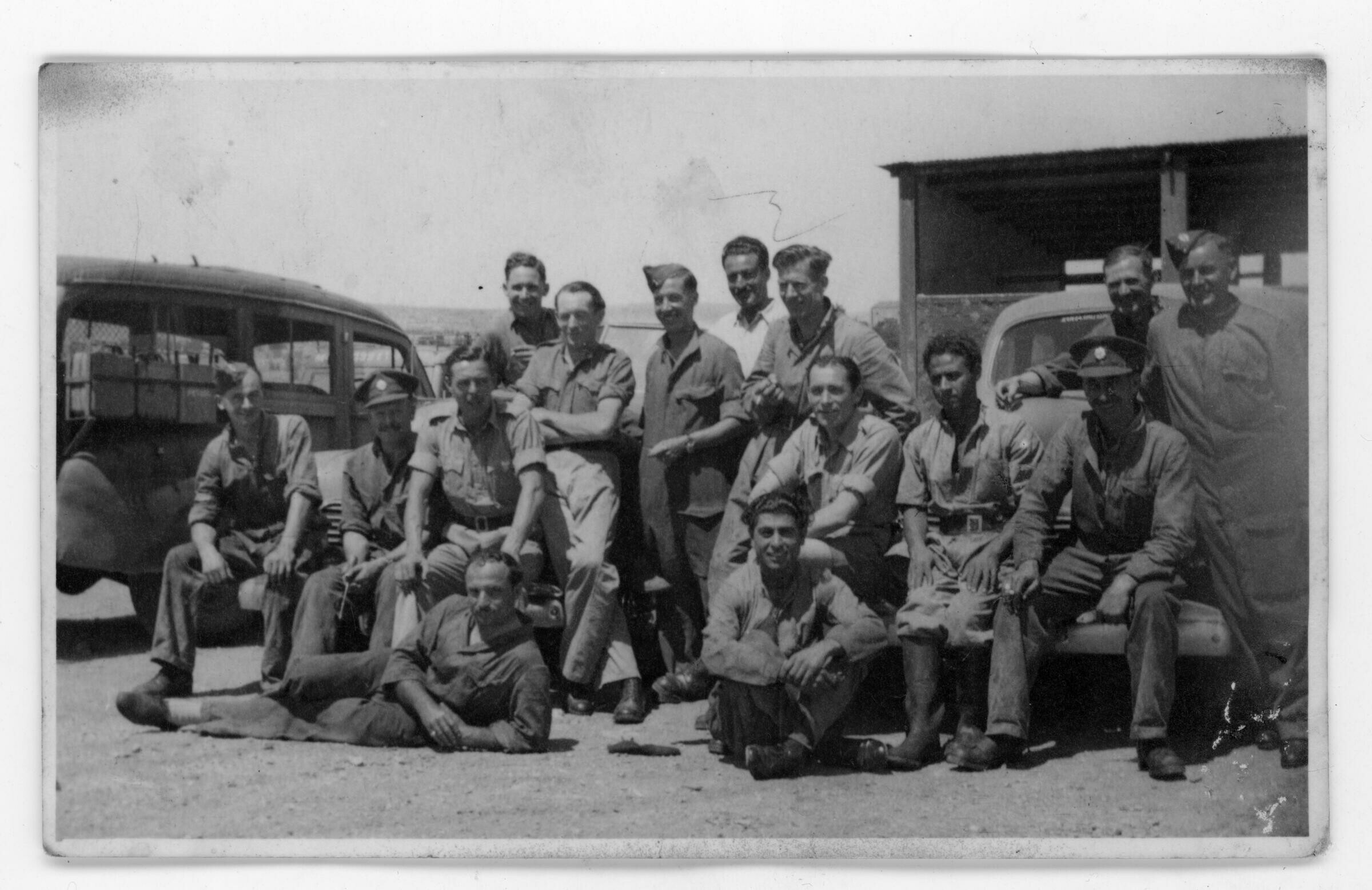 Retrato grupal de plano entero de hombres posando por delante de unos automóviles. Algunos están de pie y otros están sentados o recostados en el suelo. Lleva puesto el uniforme de una institución/agrupación no identificada.