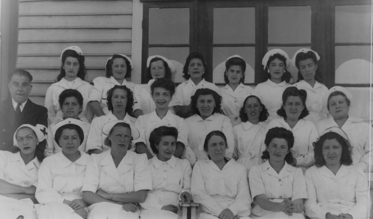 Retrato grupal de plano entero de mujeres de la cruz roja, formadas en tres hileras mientras posan en compañía de un hombre de Traje, quien está ubicado al costado izquierdo. La tercera mujer sentada de izquierda a derecha es Olga Boettcher. Llevan puesto sus uniformes y se encuentran reunidas en las afueras de una estructura no identificada.