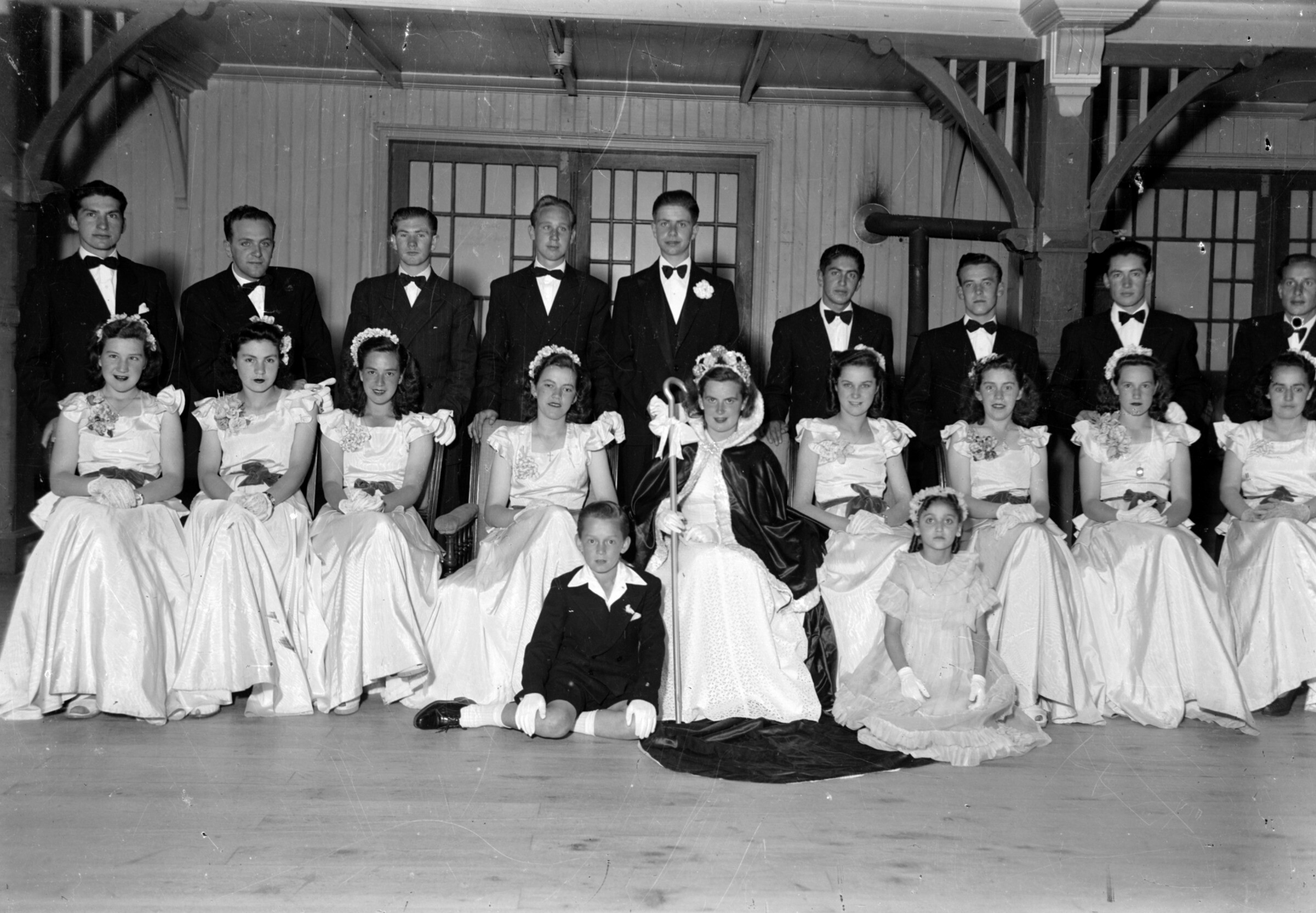Plano general de un grupo de jóvenes posando elegantemente vestidos junto a una reina en un salón