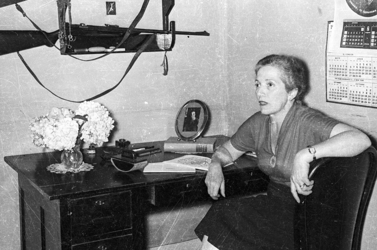 plano general de una mujer sentada de Medio perfil en una silla junto a un escritorio. Sobre el, hay un florero, un timbre, un libro, documentos y una Fotografía. también, en la pared hay un rifle colgando y un calendario colgado. Se encuentra en el interior de una infraestructura no identificada