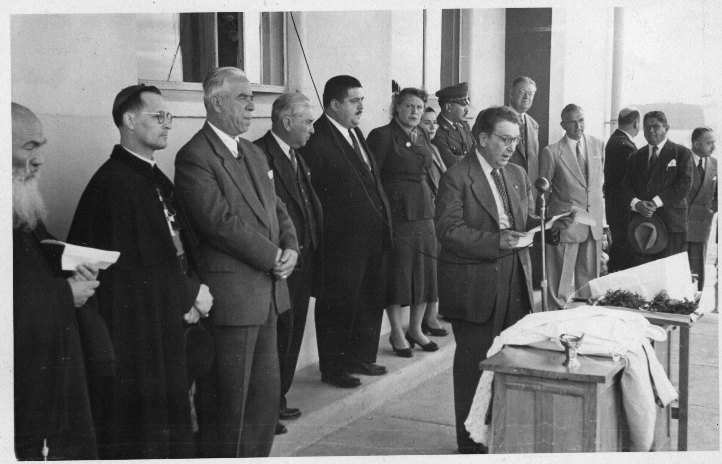 plano general de un hombre leyendo un documento frente a un micrófono. A sus espaldas, formados en una hilera, se encuentra dos sacerdotes, tres hombres, Olga Boettcher, una mujer, un carabinero y otro grupo de hombres. Todos le acompañan en la inauguración del hospital Dr. Juan Morey de La Unión realizado en el frontis del edificio