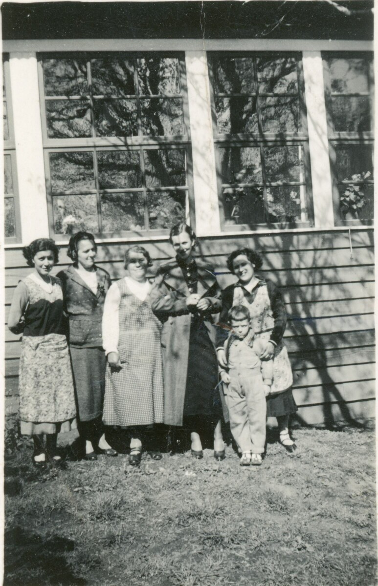 Retrato grupal de plano entero de mujeres y un niño posando junto a una casa con ventanales. Lugar no identificado.